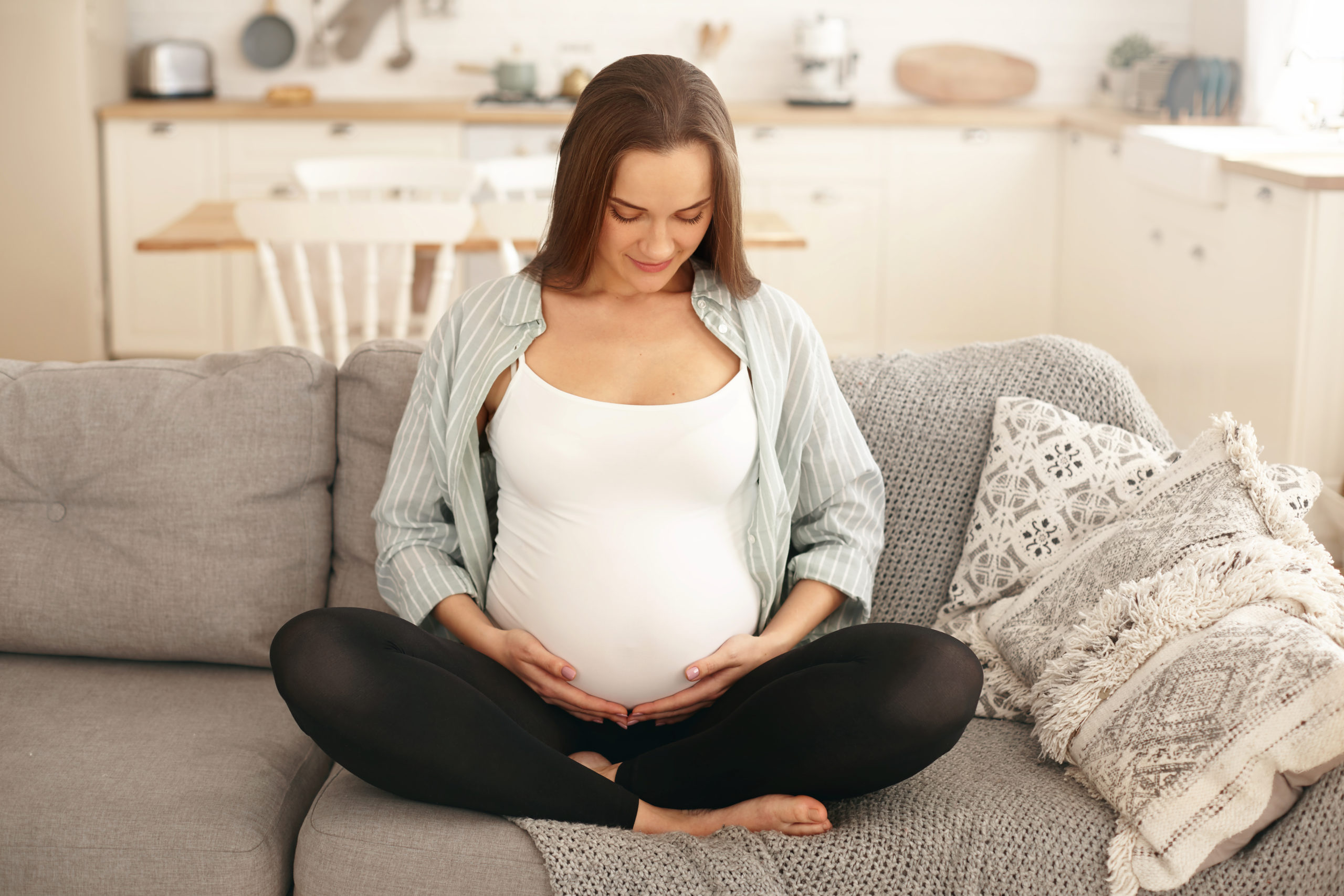 People, lifestyle, pregnancy and anticipation concept. Beautiful young brunette nin month pregnant female sitting barefooted on couch in living room, holding hands on her tummy, feeling quickening